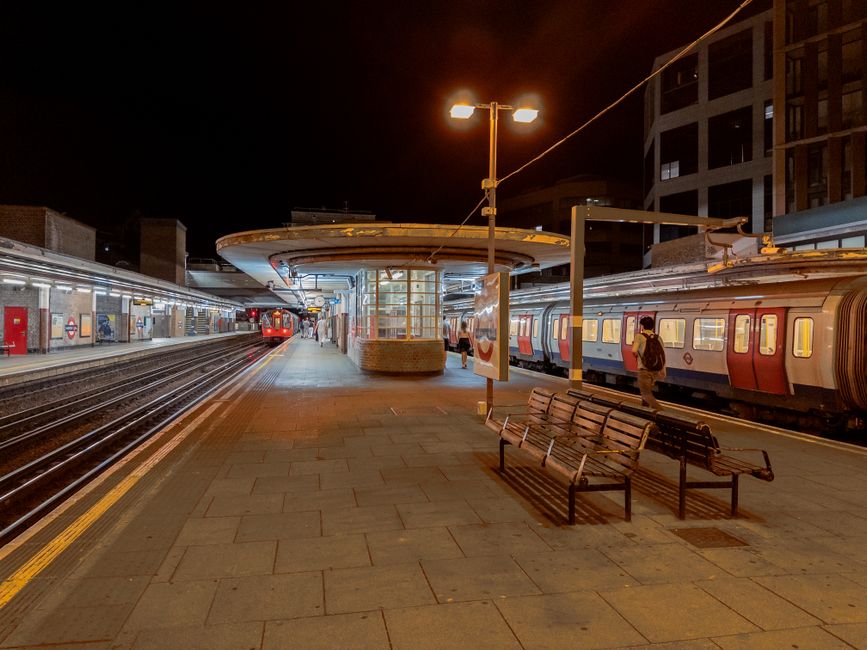 A nighttime scene at Wimbledon railway station showing a platform with a row of benches in the foreground, made of wooden slats with metal frames, positioned on a paved surface. Behind the benches, a 