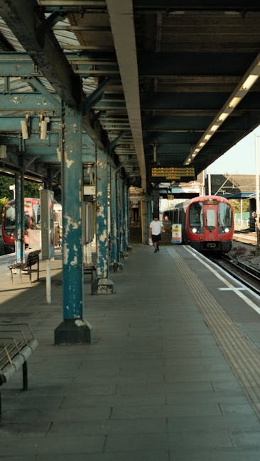 A nighttime scene at Wimbledon railway station showing a platform with a row of benches in the foreground, made of wooden slats with metal frames, positioned on a paved surface. Behind the benches, a 