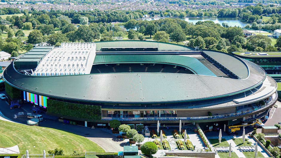 Aerial view of a modern, oval-shaped stadium with a curved green roofing structure and a partially covered outdoor seating area. The stadium is surrounded by landscaped grounds featuring neatly trimmed lawns, arranged patio tables with umbrellas, and decorative shrubs. Adjacent to the stadium, there is a parking area with several vehicles, including vans and cars, which are used for loading or unloading items during a house removal process. In the background, there are abundant trees, a river, and distant woodland, indicating a scenic location in a green environment. The lighting is bright and clear, suggesting a sunny day, and the image captures the scale of the stadium along with the surrounding outdoor space, which could be relevant for logistics planning in a home relocation or furniture transport context. Man with Van Wimbledon specializes in removals and moving services, and the image reflects the logistical considerations involved in loading equipment and furniture for relocation or event setup.